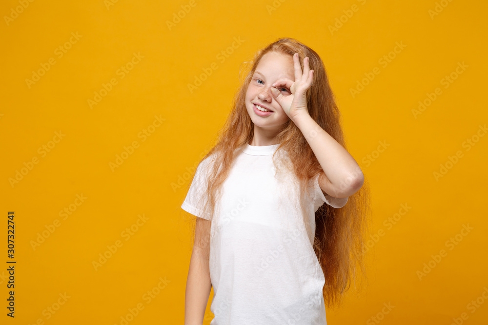 Pretty little ginger kid girl 12-13 years old in white t-shirt isolated on yellow background. Childhood lifestyle concept. Mock up copy space. Holding hand near eye, imitating binoculars or glasses. Pretty little ginger kid girl 12-13 years old in white t-shirt isolated on yellow background. Childhood lifestyle concept. Mock up copy space. Holding hand near eye, imitating binoculars or glasses.