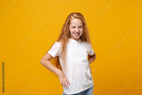 Charming little ginger kid girl 12-13 years old in white t-shirt isolated on yellow background children portrait. Childhood lifestyle concept. Mock up copy space. Standing with arms akimbo on waist.