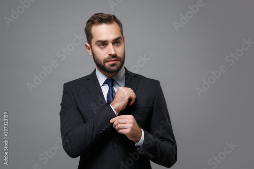 Confident young business man in classic black suit shirt tie posing isolated on grey background. Achievement career wealth business concept. Mock up copy space. Straightening sleeves, looking aside.