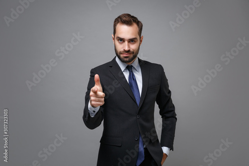 Successful young business man in classic black suit shirt tie posing isolated on grey wall background. Achievement career wealth business concept. Mock up copy space. Pointing index finger on camera.