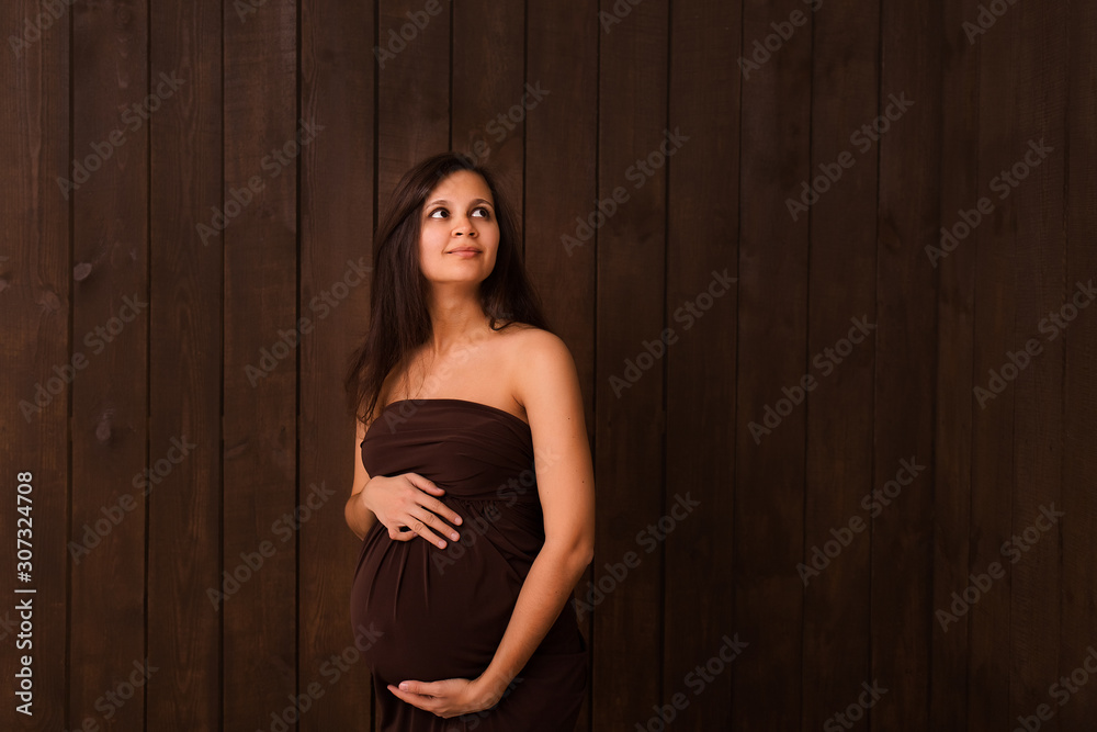 Fototapeta premium Pregnant woman in brown dress holds hands on belly on a dark brown background. Pregnancy, maternity, expectation concept. Beautiful tender mood photo of pregnancy
