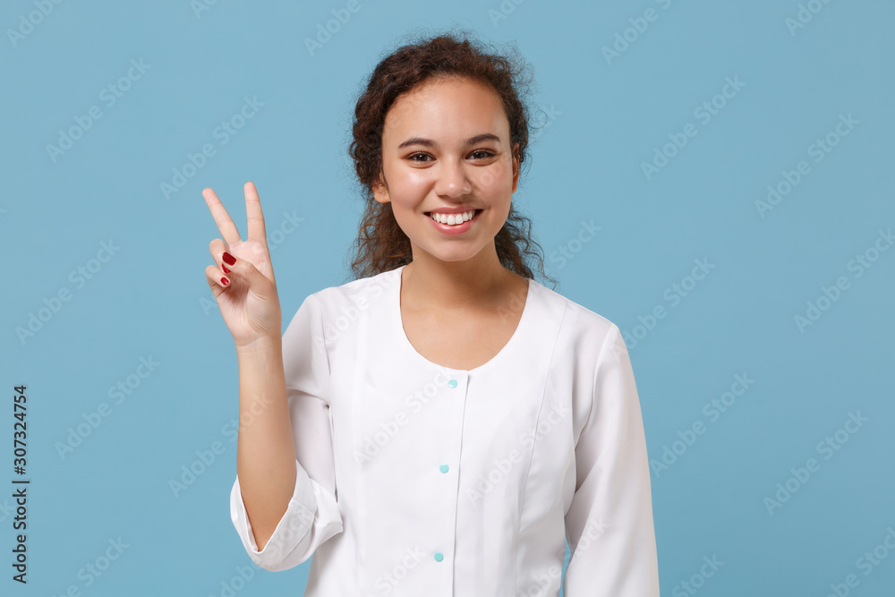 Pretty african american doctor woman isolated on blue background in studio. Female doctor in white medical gown showing victory sign. Healthcare personnel health medicine concept. Mock up copy space. Pretty african american doctor woman isolated on blue background in studio. Female doctor in white medical gown showing victory sign. Healthcare personnel health medicine concept. Mock up copy space.
