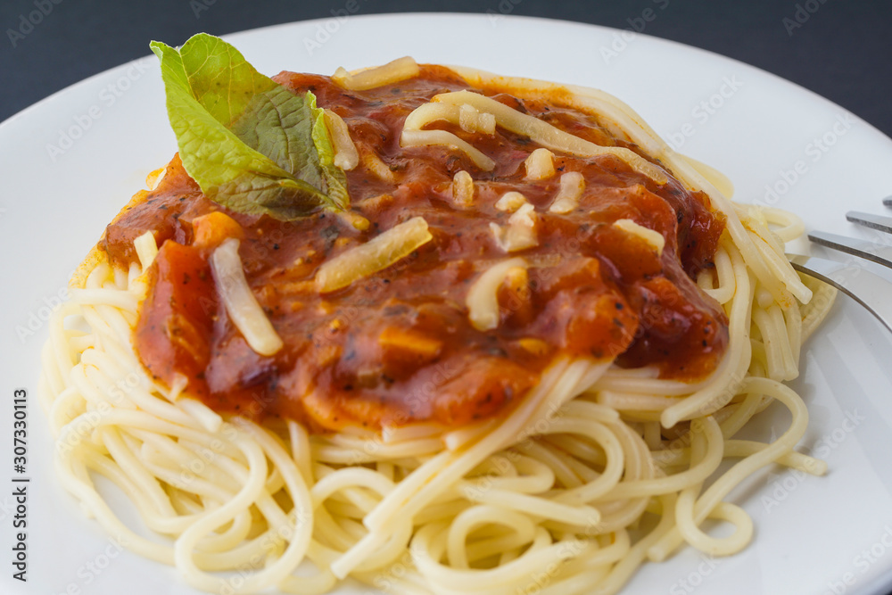 View of spagetti bolognese with green basil on top over black background. Silver fork placed next to the white plate. View of spagetti bolognese with green basil on top over black background. Silver fork placed next to the white plate.