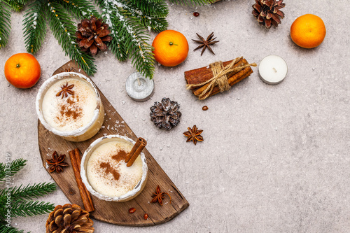 Eggnog with cinnamon and nutmeg for Christmas and winter holidays. Homemade beverage in glasses with spicy rim. Tangerines, candles, gift. Stone concrete background