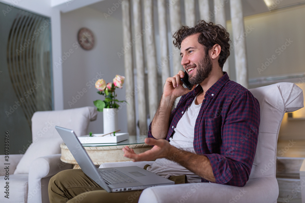 Young attractive smiling guy is browsing at his laptop and talking oh mobile phone, sitting at home on the cozy sofa, wearing casual outfit. Freelance business work from home concept Young attractive smiling guy is browsing at his laptop and talking oh mobile phone, sitting at home on the cozy sofa, wearing casual outfit. Freelance business work from home concept