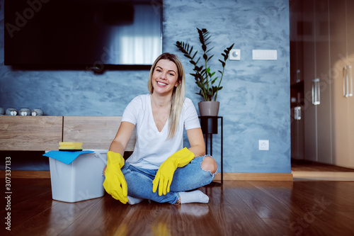 Young attractive caucasian smiling blonde housewife with rubber gloves on hands sitting on the floor with legs crossed and looking at camera. Next to her is bucket.