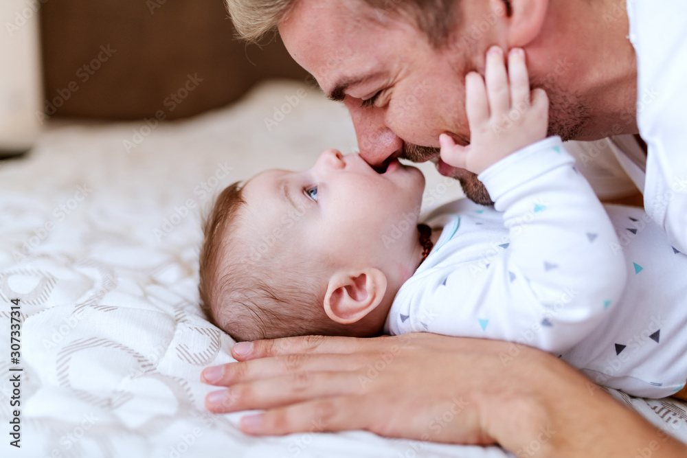 Playful adorable caucasian baby boy six months old lying in bed and biting his dad's nose. Father cuddling baby and laughing. Playful adorable caucasian baby boy six months old lying in bed and biting his dad's nose. Father cuddling baby and laughing.
