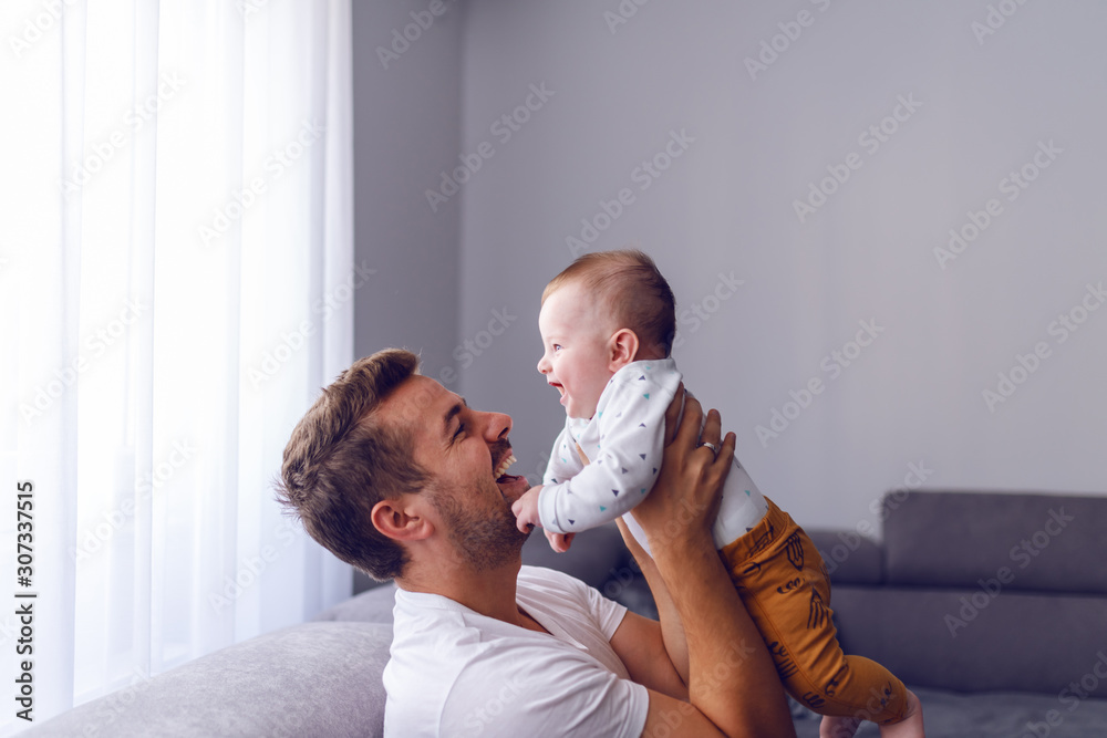 Playful handsome caucasian young dad lifting his loving 6 months old baby boy while sitting on sofa in living room. Baby enjoying time with father and laughing. - obrazy, fototapety, plakaty 