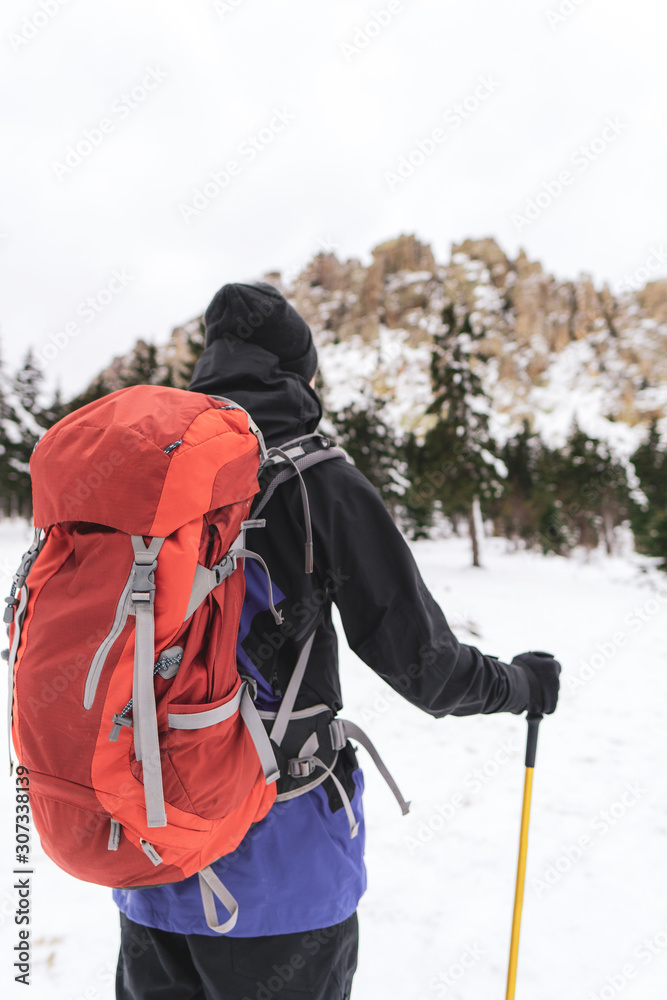 Young hiker man with backpack and trekking poles stands in winter forest and enjoys mountain range view with snowy steep stony slopes; winter outdoor activity, sports lifestyle; man traveling alone