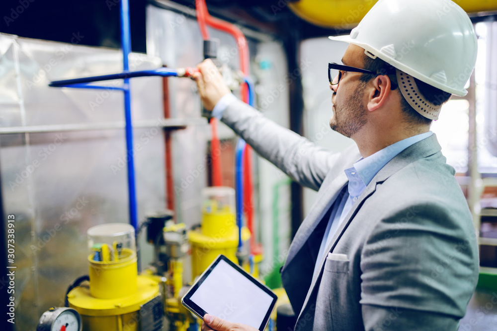 Handsome caucasian supervisor in suit and with helmet on head holding tablet and checking on boiler in factory. Handsome caucasian supervisor in suit and with helmet on head holding tablet and checking on boiler in factory.
