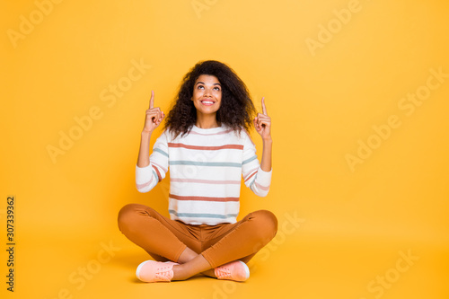 Photo of attractive smart clever confident hipster asking you to look over her head pointing finger on empty space wearing striped pullover casual pants sit floor isolated background