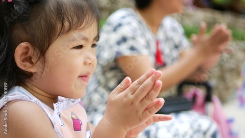 Little Asian baby girl, 26 months old, smiling and clapping her hands while enjoys watching an outdoor show with her mother