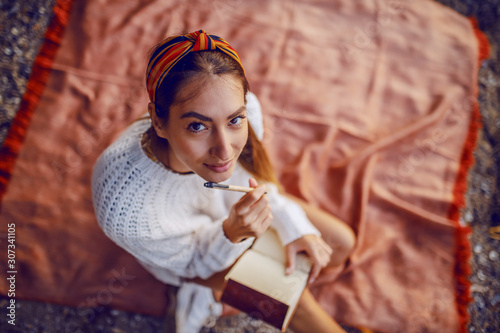 Murais de parede Top view of attractive caucasian brunette in sweater and with headband sitting on blanket outdoors and holding pen and diary in hands while looking at camera