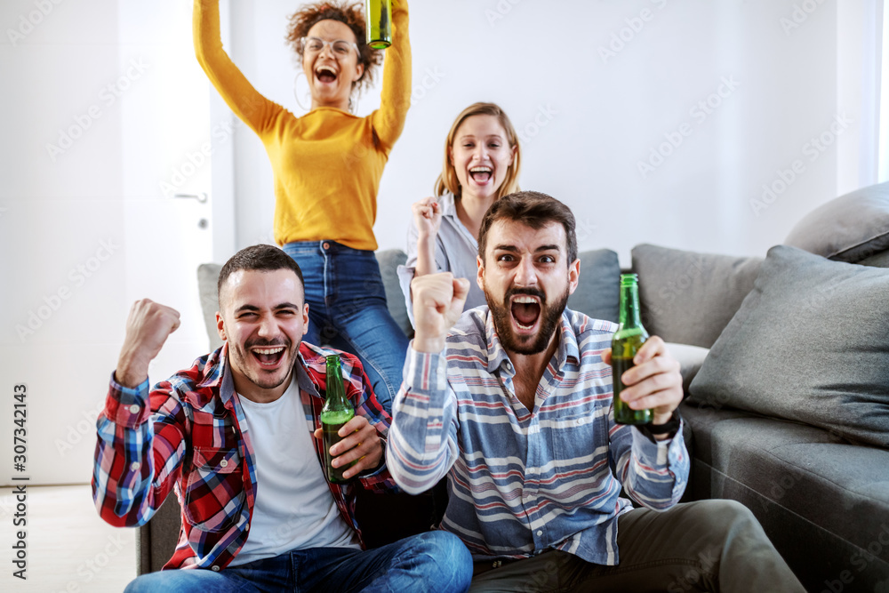 Group of friend sitting in living room and cheering for their favorite football team. They all holding beer bottles.