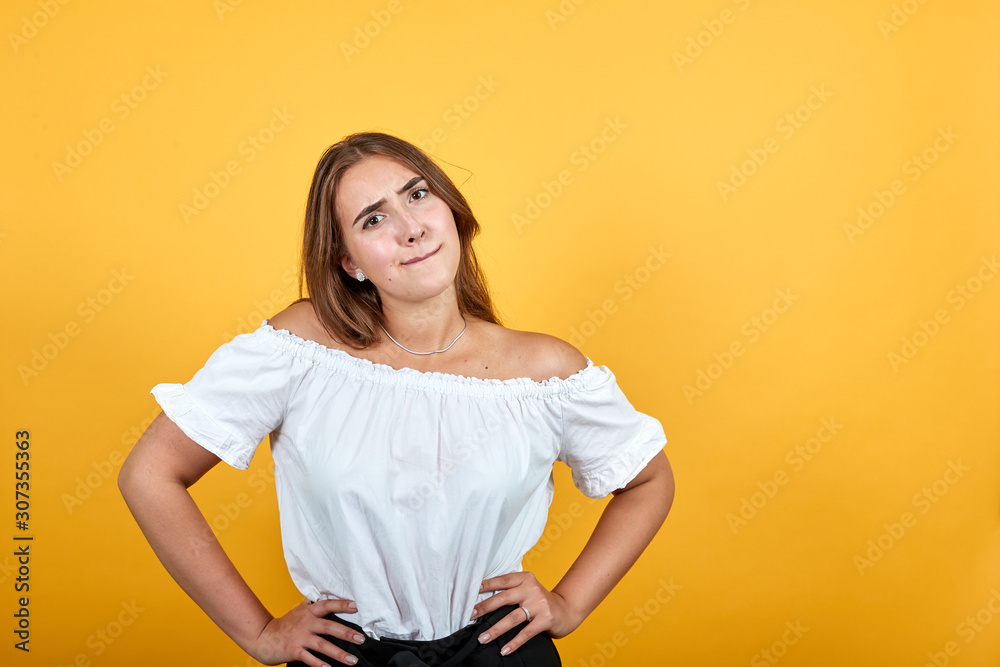 Confused young woman keeping hand on waist, looking disappointed isolated on orange background in studio in casual white shirt. People sincere emotions, lifestyle concept. Confused young woman keeping hand on waist, looking disappointed isolated on orange background in studio in casual white shirt. People sincere emotions, lifestyle concept.