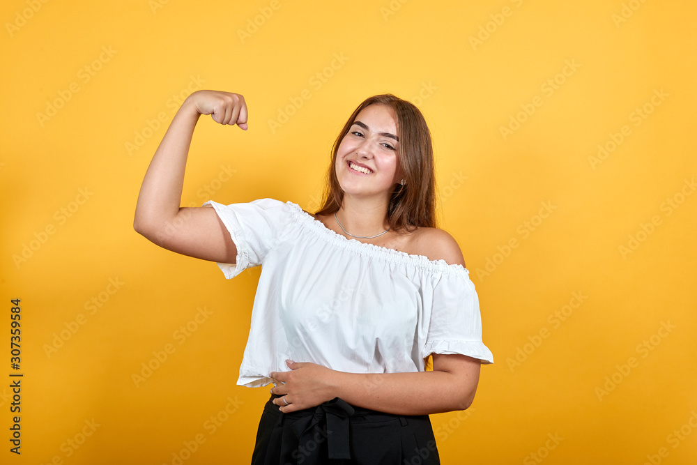 Strong charming young woman showing muscules, smiling isolated on orange background in studio in casual white shirt. People sincere emotions, lifestyle concept. Strong charming young woman showing muscules, smiling isolated on orange background in studio in casual white shirt. People sincere emotions, lifestyle concept.