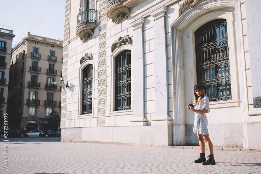 Full length portrait of cheerful female tourist dressed in casual apparel smiling at camera during sightseeing time around Barcelona, positive hipster girl with modern camera equipment in hand Full length portrait of cheerful female tourist dressed in casual apparel smiling at camera during sightseeing time around Barcelona, positive hipster girl with modern camera equipment in hand