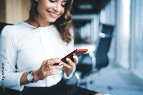 Cheerful young beautiful woman in formal outfit sitting surfing smartphone in workplace