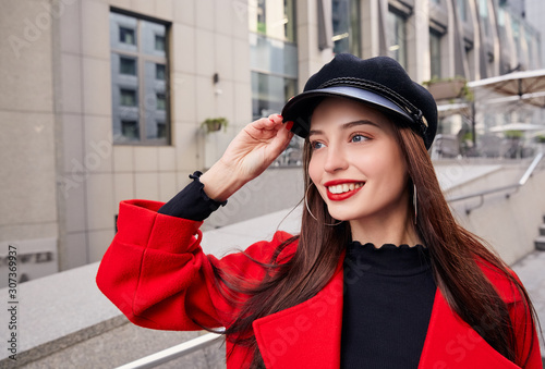 Ravishing luscious delightsome girl in red overcoat holds her kepi by hand, looks into the distance and smiles