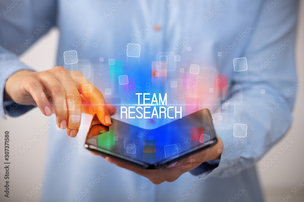 Young man holding a foldable smartphone with TEAM RESEARCH inscription, educational concept Young man holding a foldable smartphone with TEAM RESEARCH inscription, educational concept