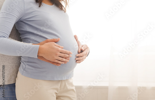 Loving husband embracing pregnant wife from behind near window at home