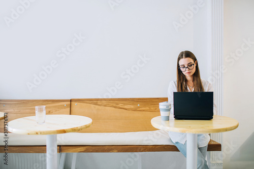 Young concentrated woman working on laptop in a coffee shop, a cup of coffee beside. Girl sitting at the table with black laptop. Copy space.