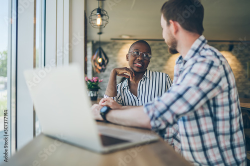 Smart man speaking to black woman at table with laptop