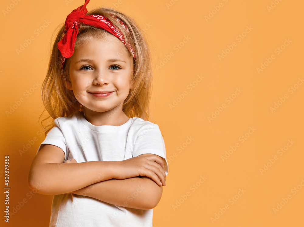 Kid girl in white t-shirt and red headband is standing at copy space with her arms crossed, looking at us on yellow  - obrazy, fototapety, plakaty 