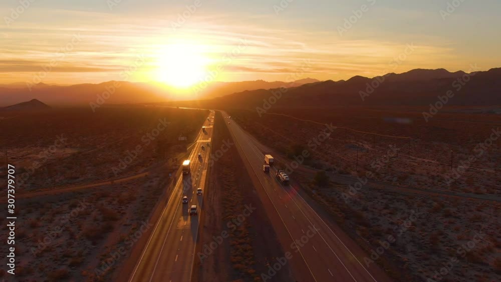 AERIAL, LENS FLARE: Scenic shot of 18 wheeler trucks and cars crossing Mojave desert at dusk. Golden evening sun rays shine on the traffic moving up and down the straight freeway in rural California.