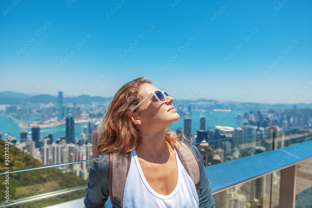 Beautiful young woman smiles happily and sets her face to the sun on the background of skyscrapers in Hong Kong, traveling to Asia