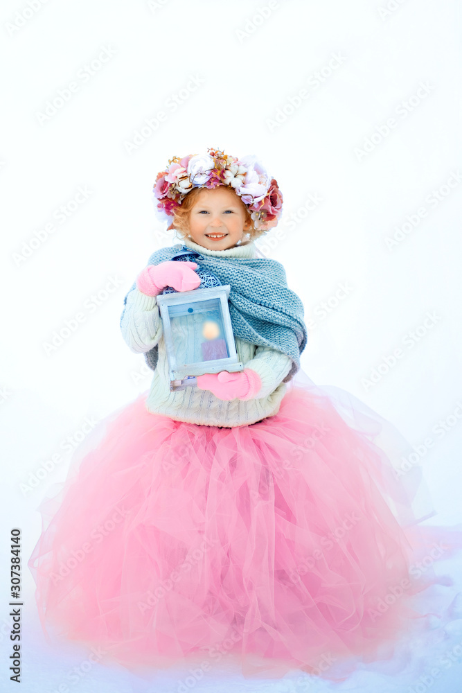 Very sweet beautiful little smiling girl child in pink skirt and mittens, white pullover, gray scarf and floral wreath with old fashioned lantern in hands. Snow is around. Space for text
