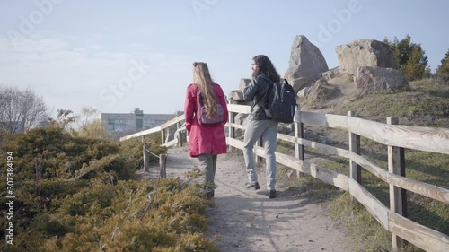 Camera following Middle Eastern man and Caucasian woman walking along wooden fence outdoors. Positive hippie couple dating in autumn park. Togetherness, leisure, resting. photo