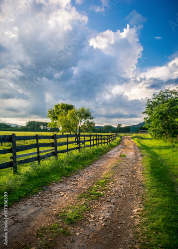 Road with a wooden fence to a green farm in the light of the rays of the sun