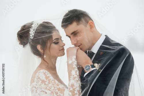 Newlyweds in love under a veil. The groom kisses the bride's hand. Wind blows veil on bride and groom looking at each other with love. Wedding photo session in the winter outdoors.