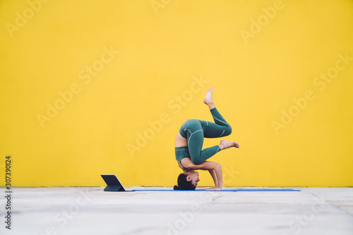 Fototapeta Young sporty fit woman exercising yoga in tripod headstand pose using tablet in