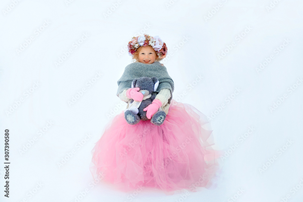 Fototapeta premium Very sweet beautiful little smiling girl child in pink skirt and mittens, white pullover, gray scarf and floral wreath with old taddy bear in hands. Snow around. Space for text