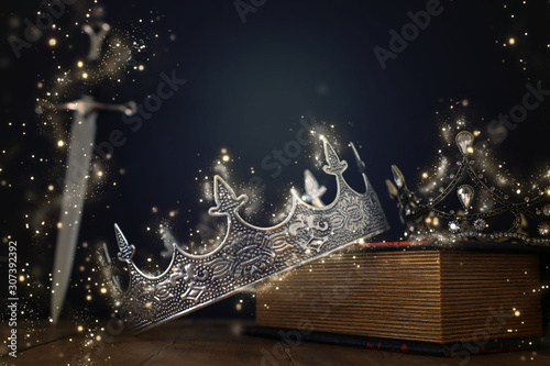low key image of beautiful queen/king crown over antique book next to sword. fantasy medieval period. Selective focus. Glitter sparkle lights