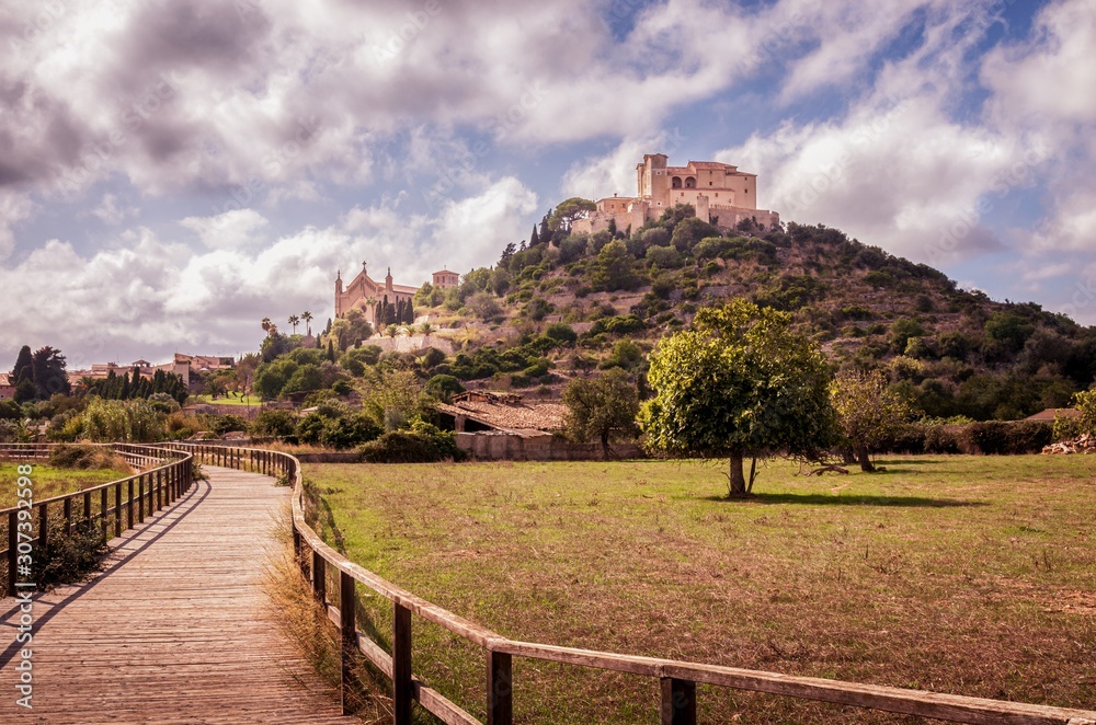 Parish church and Pilgrimage church of Sant Salvador Arta, located on a hill with fields and a wooden walkway in the foreground, daytime with blue sky and puffy white clouds, Mallorca, Spain.