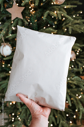 Man holds a Christmas present in hands. Happy holiday, gift box. Christmas tree with garlands in home, beautiful lights. Surprise for Boxing day, New Year and Christmas. Kraft wrapping paper and twine