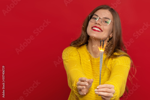 selective focus of happy girl in eyeglasses holding sparklers, wears yellow sweater, isolated on red background, expresses good emotions. Christmas concept