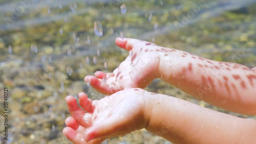 Childs hands under the drops of water with sun reflecting on the skin. Water drops falling on little hands on seashore background. Infant fingers are playing with water. Toddlers arms under the water