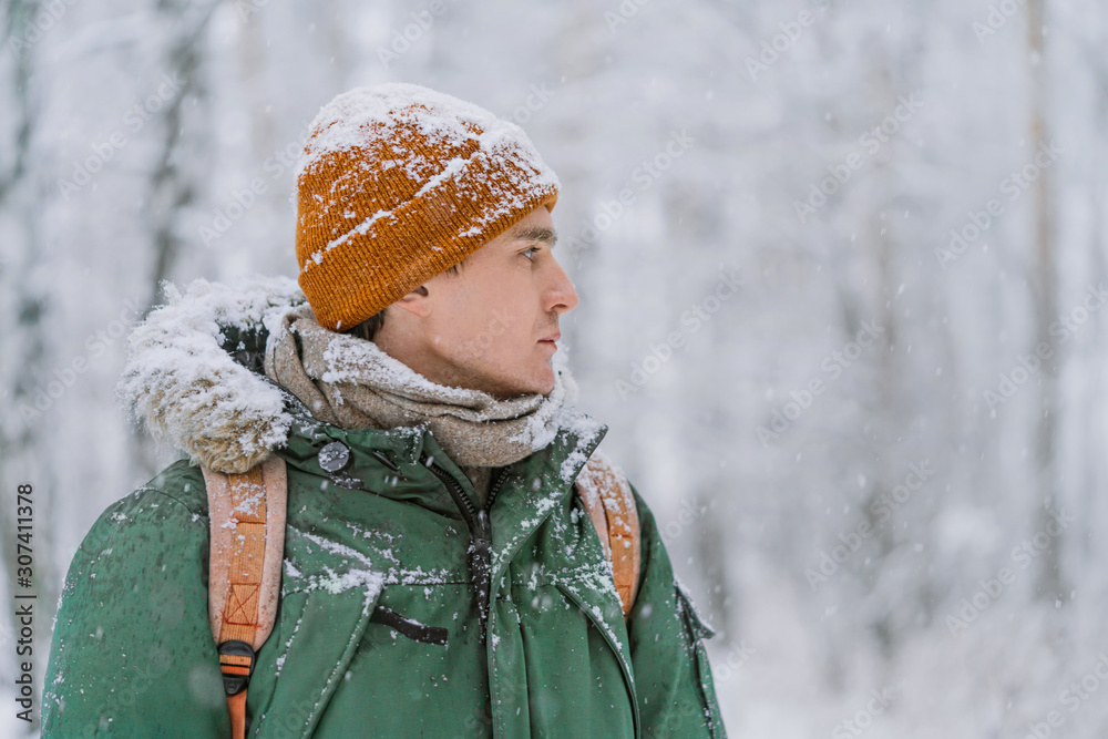 Fototapeta premium A man in clothes covered with snow looks forward to the snow-covered forest. The man is dressed in a green warm jacket and a red hat. Search in the winter forest.