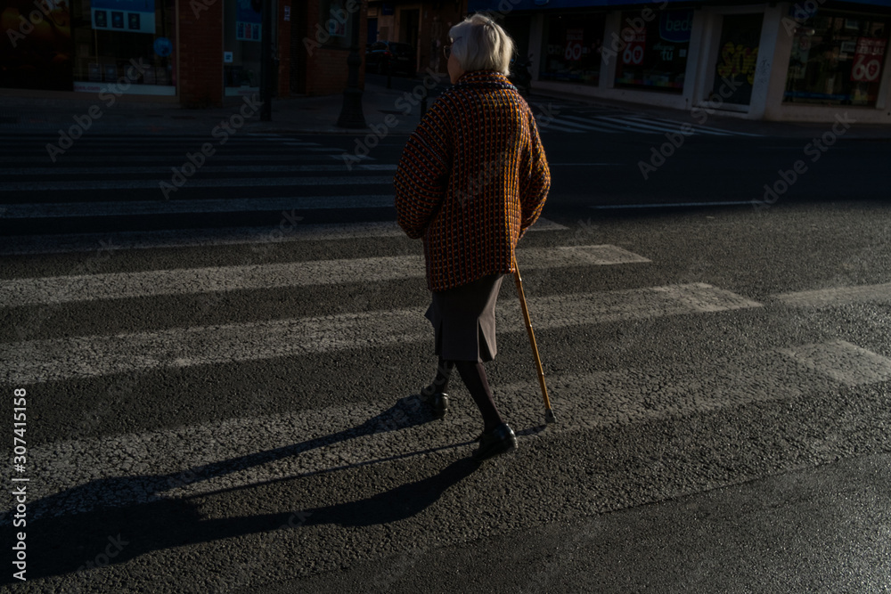 old lady with cane crossing a zebra crossing illuminated by sunset light in a city