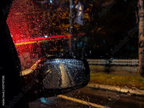 Amazing atmosphere during night while driving on a rainy street with closeup of red car street lights and drops of rain in focus on a car window glass with side rear view mirror