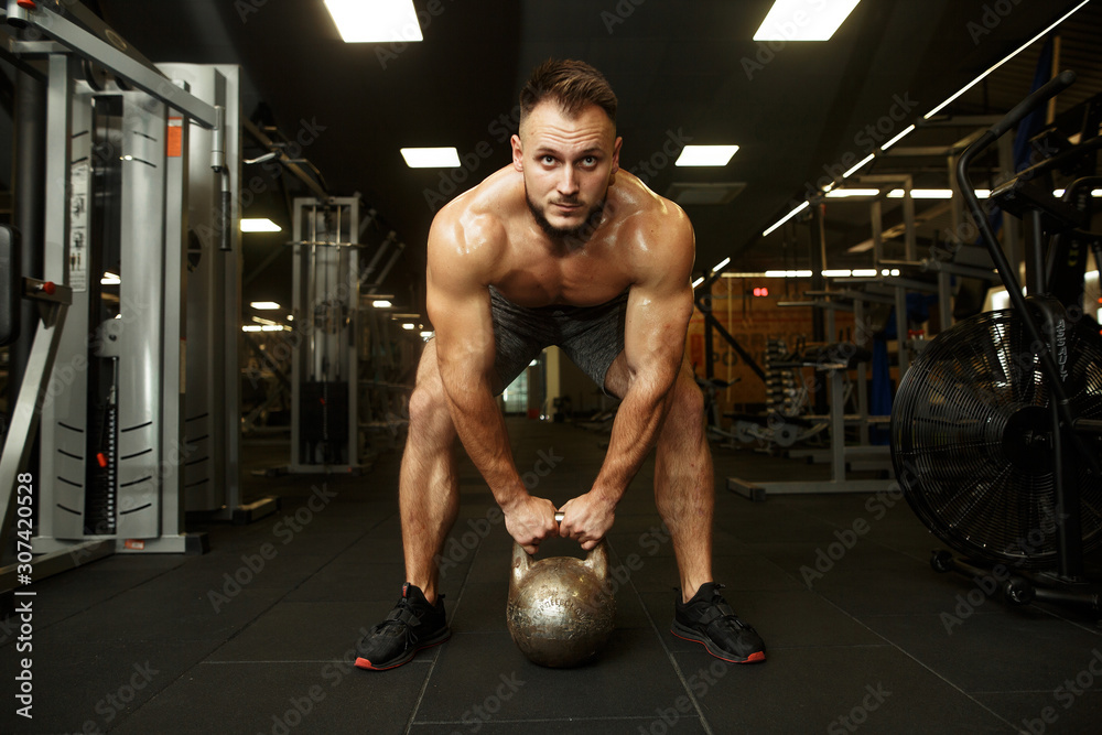 Strong handsome bearded man holding kettlebell and doing dumbbell front swings in the popular dark gym. SPORT IS LOVE. Strong handsome bearded man holding kettlebell and doing dumbbell front swings in the popular dark gym. SPORT IS LOVE.