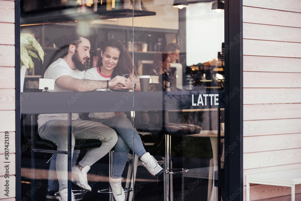 View through the glass of a young couple in love sitting in a cafe and drinking coffee and watching a video about traveling on a smartphone using high-speed free Internet. Travel dream concept