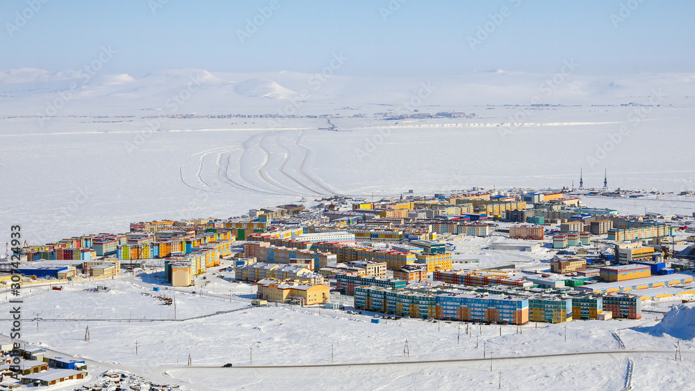 Winter aerial panorama of the city of Anadyr. Top view of the colorful buildings and the snowy expanses of the Arctic. In the distance, an ice crossing through the Anadyr estuary. Chukotka, Russia.