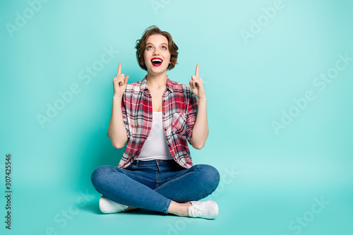 Full length photo of amazed funky crazy girl sit legs floor crossed point attention index finger copy space indicate ads promo scream wow omg wear denim jeans isolated teal color background © deagreez