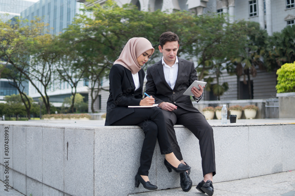 Asian woman and Caucasian man sitting in discussion while holding electronic tablet and writing a note. Asian woman and Caucasian man sitting in discussion while holding electronic tablet and writing a note.