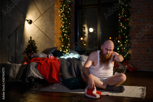 Humorous image of a man without pants in front of Christmas. Bald Santa Claus with a red beard unpacks a gift. A parody of a glamorous women's New Year photo shoot. Joke.